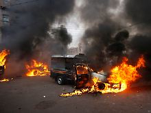 Nach dem Anschlag kommt es zu Straßenschlachten; auch Polizeiautos gehen in Flammen auf.