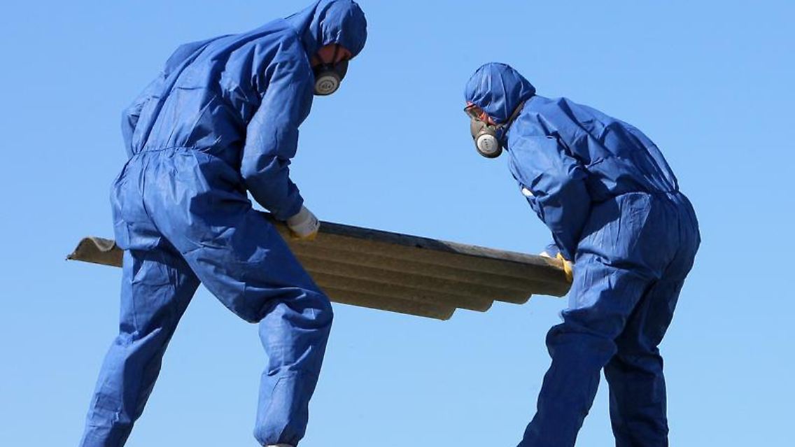 Nur für Profis: Arbeiter in Schutzanzügen und mit Luftfiltern über Mund und Nase entfernen Asbestplatten vom Dach eines alten Garagenkomplexes.