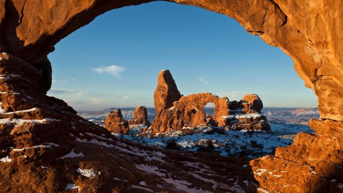 Der Turret Arch und der North Window Bogen, hier im Bild, sind zwei der massiven Steinbögen in der sogenannten Windows Section des Arches Nationalparks. Die Felsformationen sind besonders bei Sonnenauf- und Untergang außergewöhnlich fotogen: Das Sonnenlicht lässt das Gestein dann noch röter erscheinen.