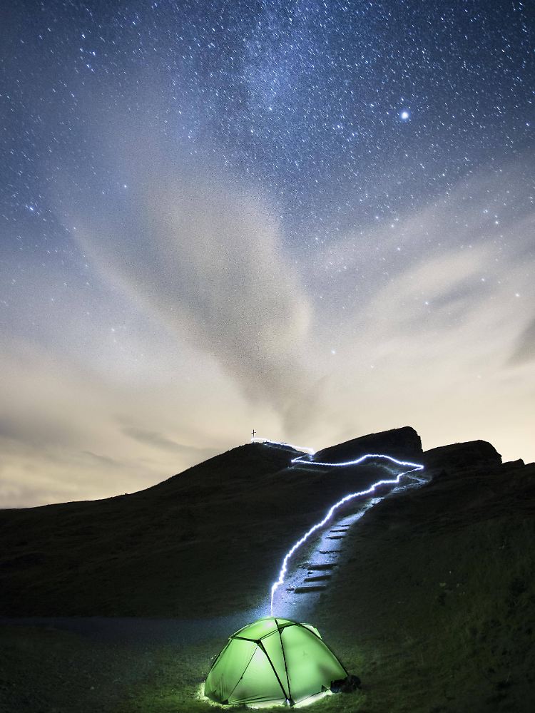 Zeit für nächtliche Beobachtungen: Grünes leuchtendes Zelt unter der Milchstraße mit Lichtspuren einer Taschenlampe.