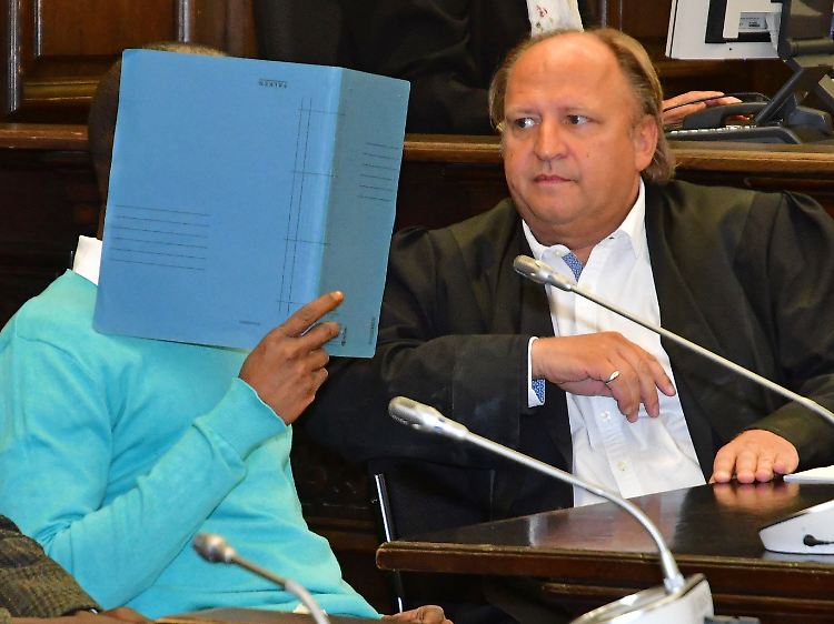 Der Angeklagte sitzt im Gerichtssaal neben seinem Anwalt Tim Burkert (r.) und verbirgt sein Gesicht mit einem Aktendeckel.