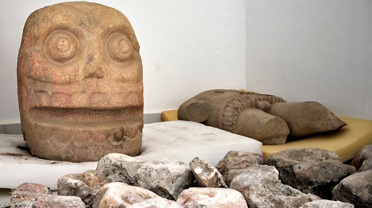 Die Aufnahme zeigt eine Schädelskulptur aus Stein und eine Statue, die die Gottheit Xipe Tótec darstellen soll.