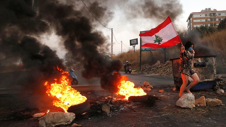 In Beirut geht eine Teilnehmerin einer Demonstration mit einer Libonon-Fahne und Mundschutz an brennenden Reifen vorbei.