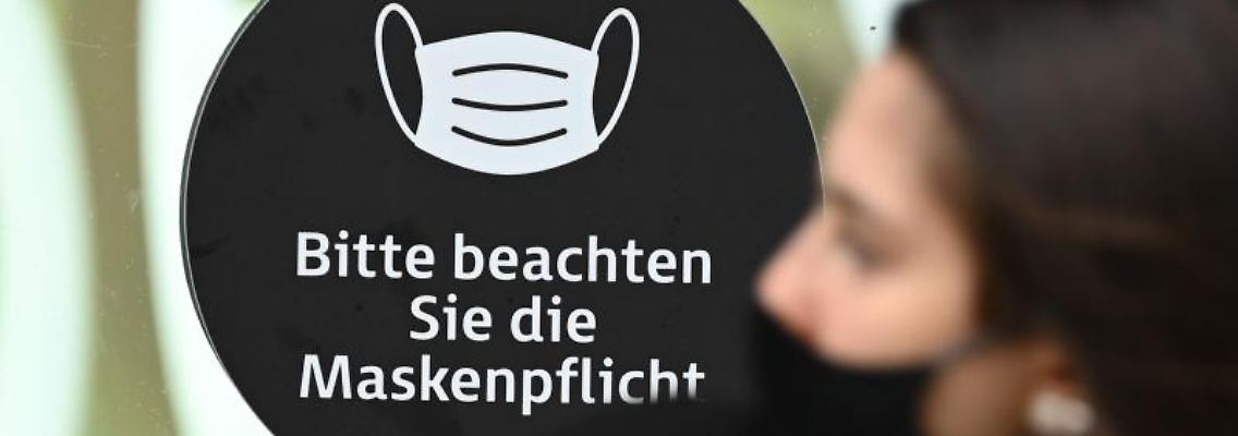 Eine Frau, die einen Mund-Nasen-Schutz trägt, an einem Hinweis mit der Aufschrift "Bitte beachten Sie die Maskenpflicht" vorbei.jpg