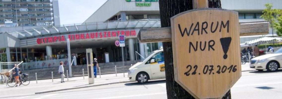 Ein Holzkreuz mit einem Schild und der Aufschrift "Warum nur! - 22.07.2016" hängt vor dem Eingang zum Olympia-Einkaufszentrum (OEZ). Foto: Peter Kneffel/dpa/Archivbild