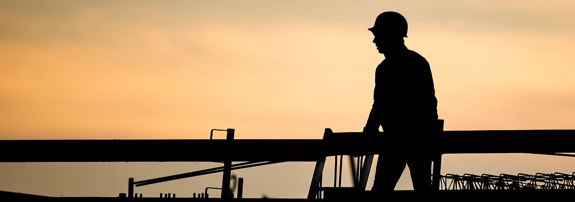Die Silhouette eines Bauarbeiters zeichnet sich vor dem verfärbten Morgenhimmel ab.