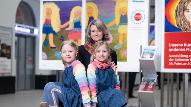 Emma (l.) und Lotte mit ihrer Mutter Katja Franke im Dresdner Hauptbahnhof in einer Ausstellung zum Tag der Seltenen Erkrankungen vor ihrem Selbstporträt.
