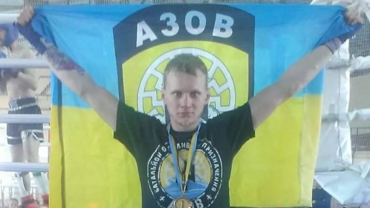 Maksym Kagal trägt eine Ukraine-Flagge mit Asow-Schriftzug.