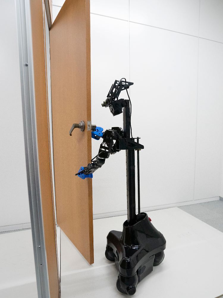 Der türöffnende Roboter hat einen Kopf mit Kamera, zwei Arme und einen Körper, der sich auf Rädern bewegt.