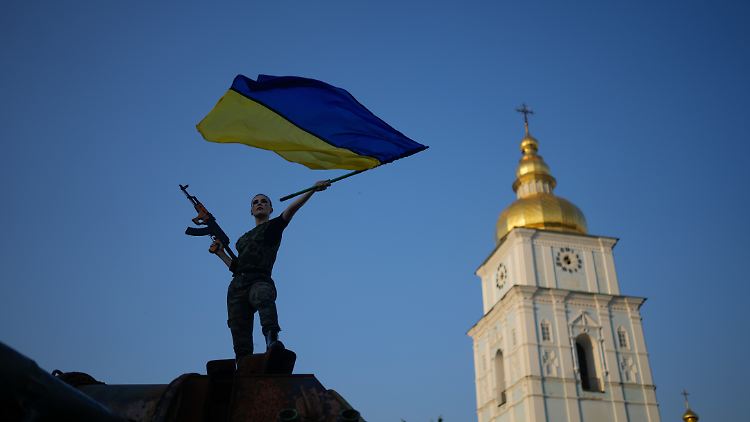 Auf einem zerstörten russischen Panzer schwenkt eine Frau in Kiew die ukrainische Flagge. 