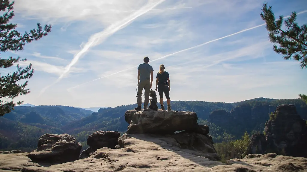 Zwei-Personen-mit-Hund-stehen-auf-einem-Felsen-in-der-Saechsischen-Schweiz