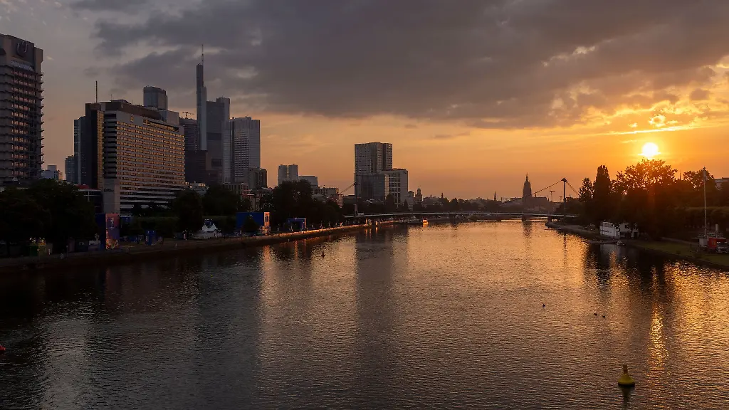 Die-Skyline-von-Frankfurt-ist-vor-der-aufgehenden-Sonne-am-Ufer-des-Mains-bei-leicht-bewoelktem-Himmel-zu-sehen