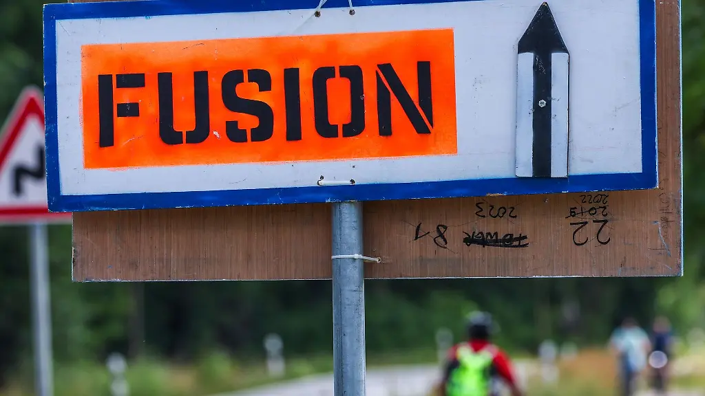 Radler-fahren-an-einem-Wegweiser-zum-Musikfestival-Fusion-an-einer-Zufahrtsstrasse-vorbei