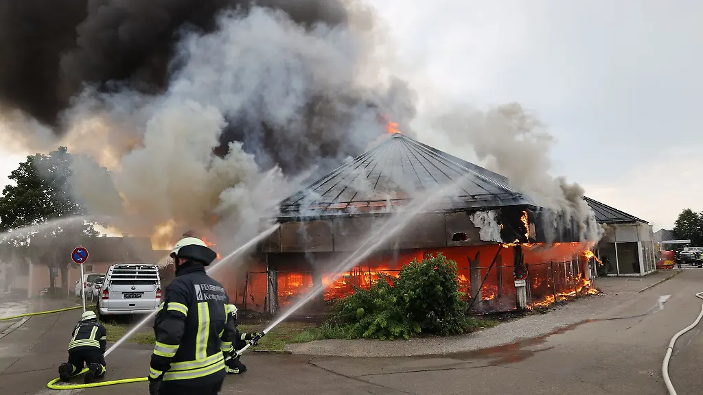 Einsatzkraefte-der-Feuerwehr-sind-bei-Loescharbeiten-vor-einem-brennenden-Gebaeude-im-Einsatz