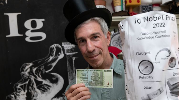 Zeremonienmeister Marc Abrahams hält einen 10-Billionen-Dollar-Schein und den Ig-Nobelpreis 2022 in seinem Büro. Der Preis wurde den Gewinnern im Voraus  zugeschickt, zusammen mit einer Anleitung zur Selbstmontage.