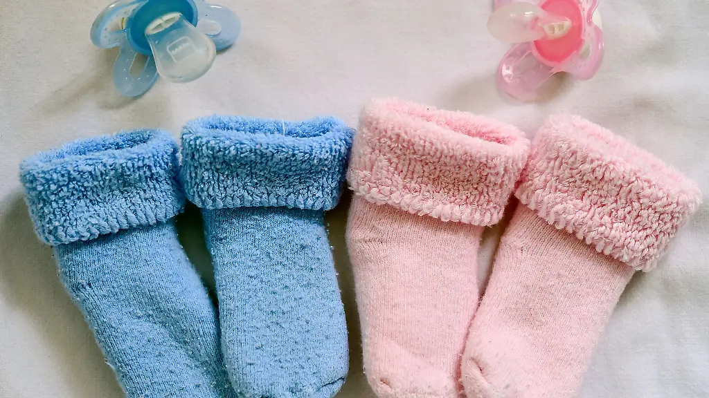Rosa-und-blaue-Soeckchen-und-dazu-die-farbigen-Nuckel-liegen-in-einem-Babybett