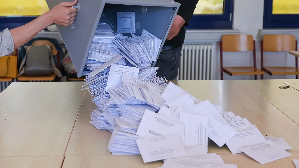 Zwei-Wahlhelfer-kippen-eine-Wahlurne-mit-den-Briefwahlstimmen-nach-der-Europawahl-in-einem-Briefwahllokal-zur-Stimmenauszaehlung-auf-einen-Tisch