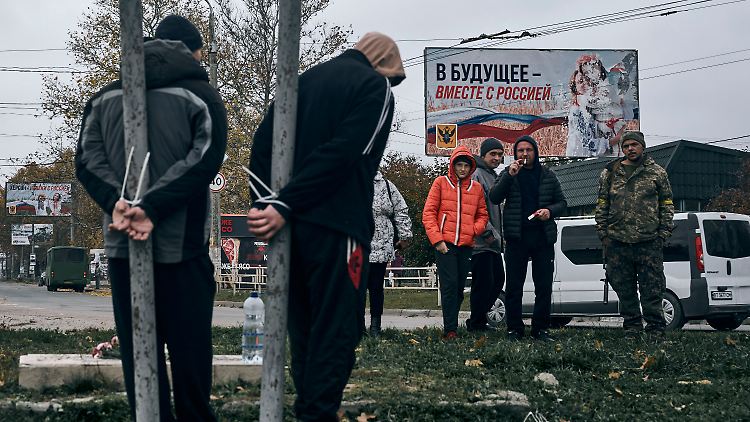 Ein ukrainischer Soldat und Anwohner stehen in Cherson neben zwei mutmaßlichen Kollaborateuren, die an Pfeiler gefesselt sind. 