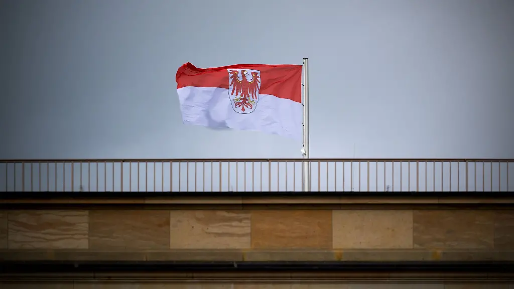Die-Flagge-des-Landes-Brandenburg-mit-dem-roten-Adler-weht-auf-dem-Landtag-vor-grauem-Himmel