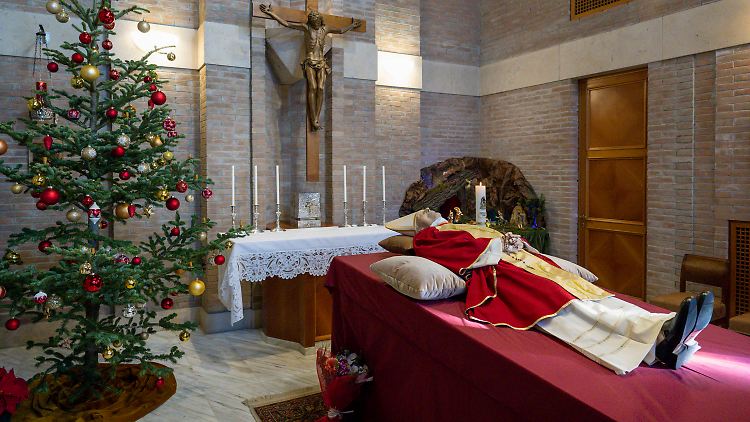 In seinen gefalteten Händen hält der verstorbene emeritierte Papst einen Rosenkranz - neben ihm stehen ein geschmückter Christbaum und eine Krippe.