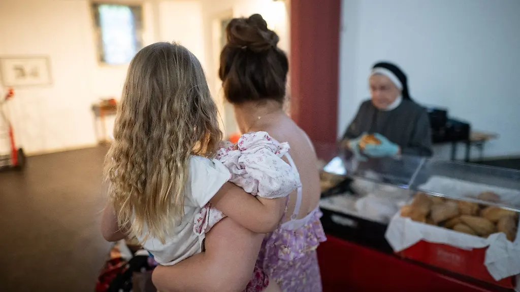 Ein-Kind-sitzt-der-Ausgabezeit-in-einer-Ausgabestelle-der-Tafel-auf-dem-Arm-ihrer-Mutter-die-Lebensmittel-abholt