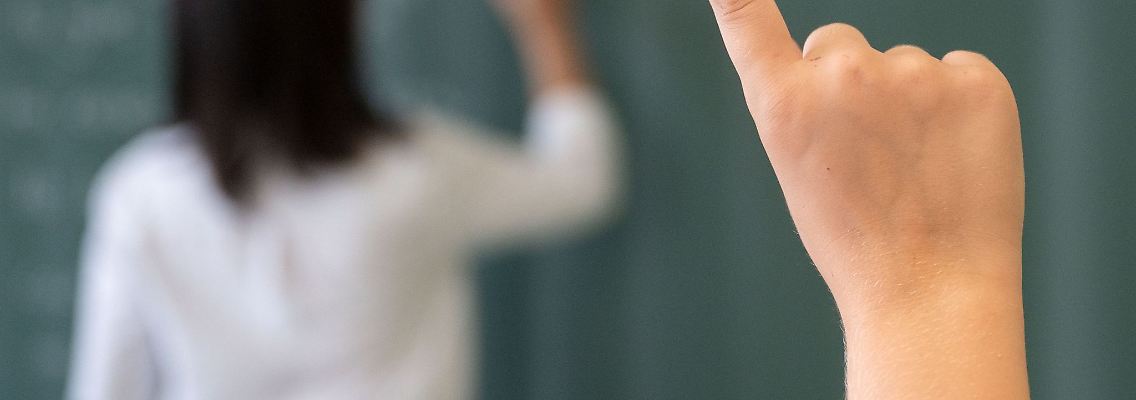 Ein Schüler meldet sich, während die Lehrerin an die Tafel schreibt.