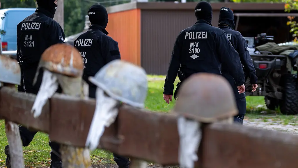 Polizisten-gehen-zur-Sicherung-von-Beweismitteln-zu-einem-Haus-vor-dem-ein-Gartenzaun-mit-rostigen-Stahlhelmen-steht