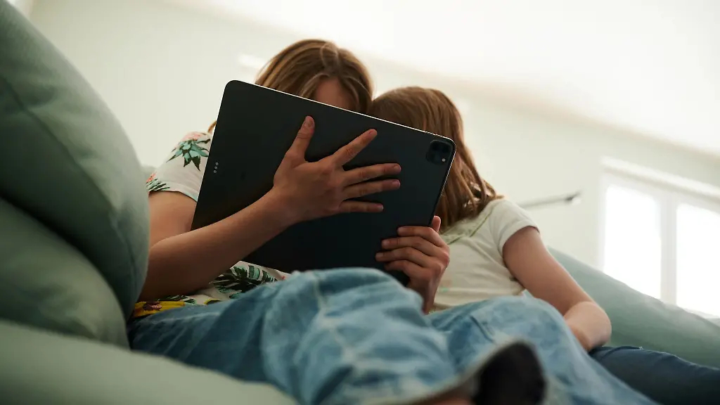Zwei-Schwestern-sitzen-auf-der-Couch-und-gucken-in-das-Tablet-ihrer-Eltern