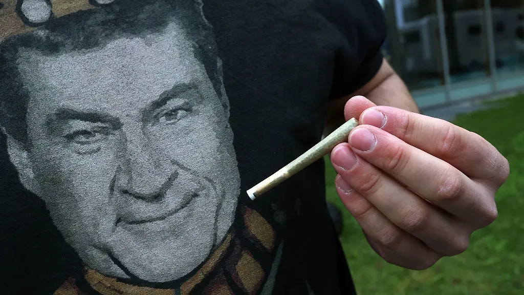 Ein-Teilnehmer-einer-Versammlung-die-unter-dem-Motto-Liberalitas-Bavariae-statt-CSU-Verbotsirrtum-angemeldet-wurde-haelt-am-Gebaeude-der-CSU-Landesleitung-vor-einem-T-Shirt-mit-einem-Portrait-des-bayerischen-Ministerpraesidenten-Markus-Soeder-CSU-einen-Joint