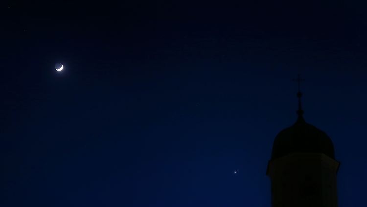 Der zunehmende Mond und der hell leuchtende Planet Venus hinter einem Kirchturm am Abendhimmel.