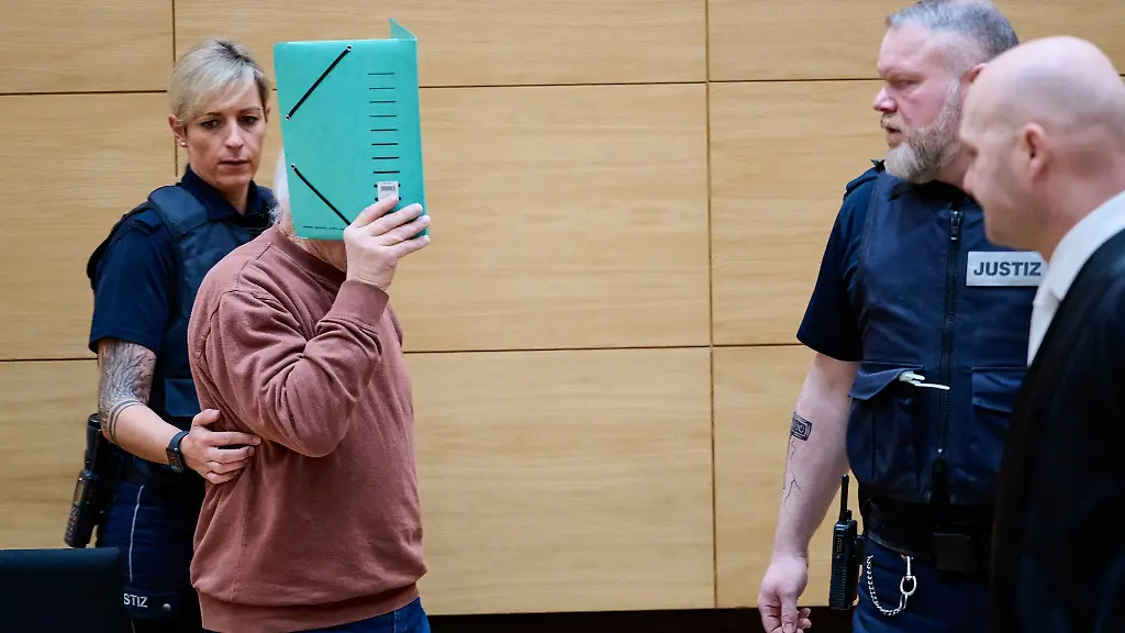 Der-Angeklagte-wird-von-einer-Justizmitarbeiterin-zu-seinem-Platz-im-Landgericht-gefuehrt-und-verbirgt-sein-Gesicht-hinter-einer-Akte