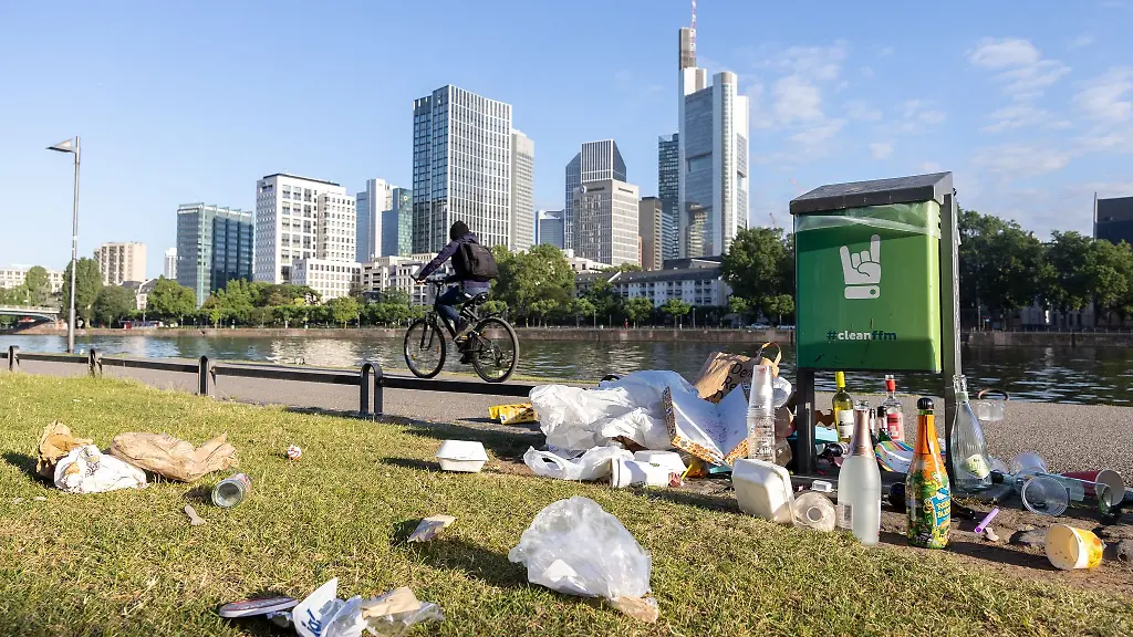 Liegengelassener-Muell-am-Mainufer-mit-der-Skyline-und-Fahrradfahrer-im-Hintergrund