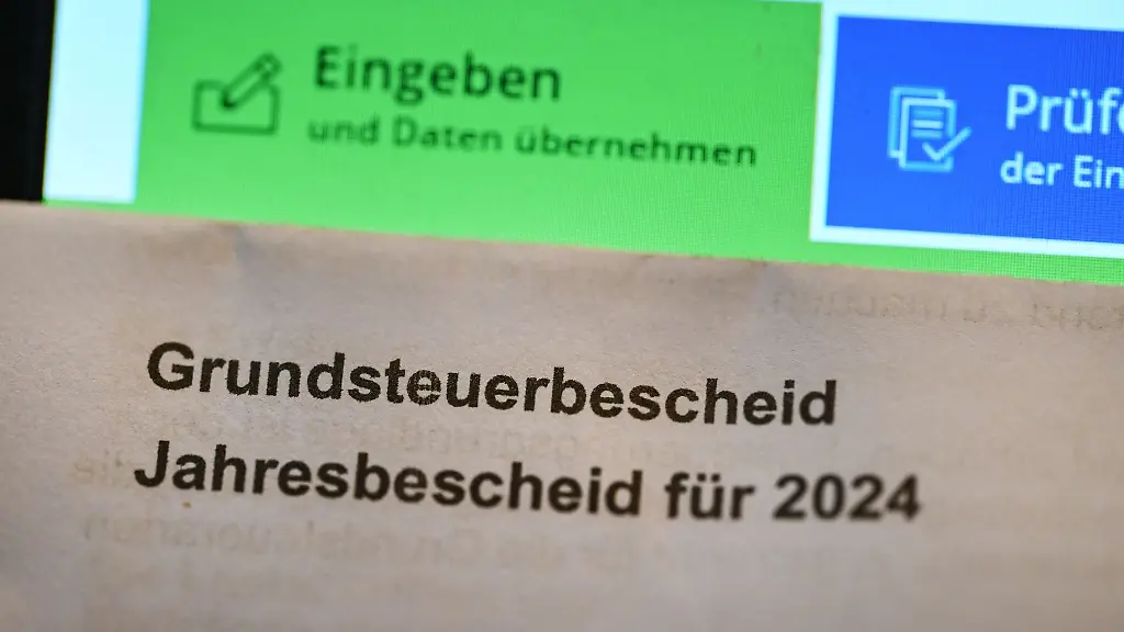 Ein-Grundsteuerbescheid-fuer-2024-wird-vor-einen-Computerbildschirm-gehalten-auf-dem-ein-Formular-der-Steuerplattform-Elster-angezeigt-wird