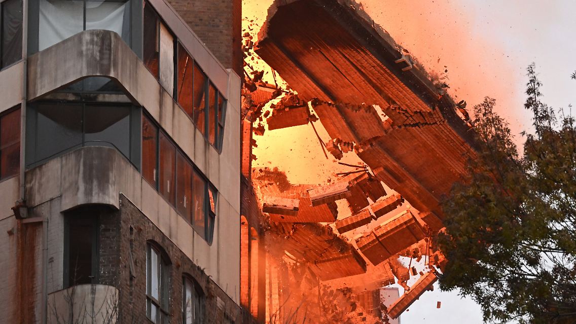 Das in Brand geratene Gebäude steht nahe an anderen Gebäuden. Trümmerteile fallen herab.