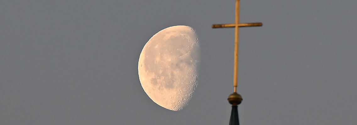 Der Mond steht im Morgenlicht hinter einem Kreuz auf einem Kirchturm.