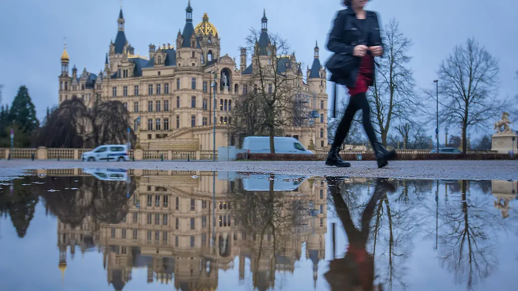 Eine-Frau-geht-an-einer-Pfuetze-vorbei-in-der-sich-das-Schweriner-Schloss-spiegelt