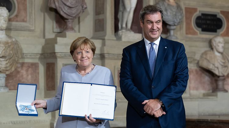 Da hat Altkanzlerin Angela Merkel die hohe Anerkennung des Freistaats Bayern schon in der Hand: So ganz ohne ein paar Spitzen lief die Verleihung des Verdienstordens aber nicht ab.