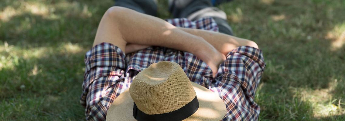 Ein Mann mit einem Sonnenhut auf dem Gesicht liegt auf einer Wiese im Schatten und ruht sich aus.