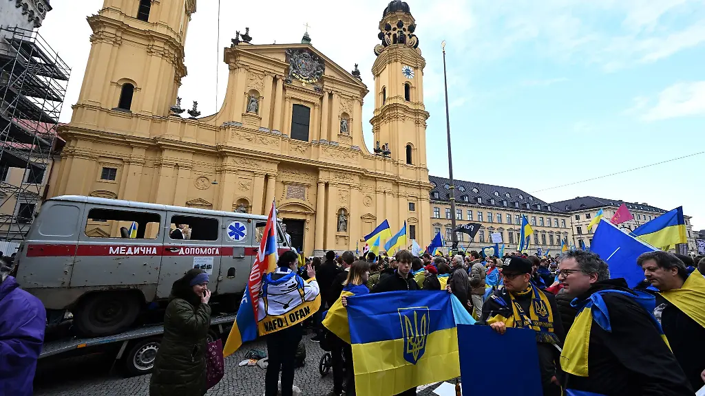 Demonstranten-stehen-mit-ukrainischen-Fahnen-vor-einem-zerschossenen-Krankenwagen-bei-einer-Demonstration-waehrend-der-Sicherheitskonferenz-auf-dem-Odeonsplatz-In-Bayern-leben-derzeit-mehr-als-160-000-Kriegsfluechtlinge-aus-der-Ukraine