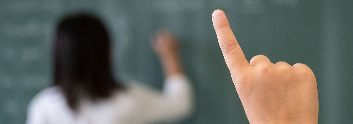 Ein Schüler meldet sich, während die Lehrerin an die Tafel schreibt.
