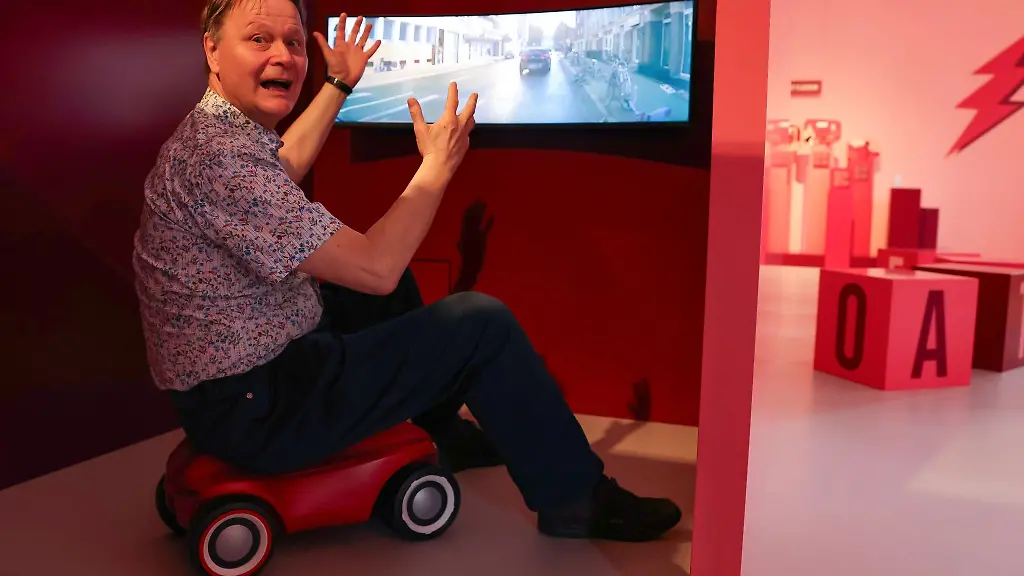 Kurator-Rolf-Bernhard-Essig-sitzt-in-der-Sonderausstellung-auf-einem-Rutschauto-und-schimpft-ueber-den-Strassenverkehr-auf-dem-Bildschirm