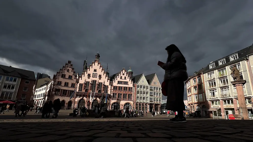 Unter-einem-dunklen-Wolkenhimmel-leuchtet-das-Rathaus-Roemer-im-Sonnenschein-auf-dem-Roemerberg-auf-dem-eine-Touristin-steht