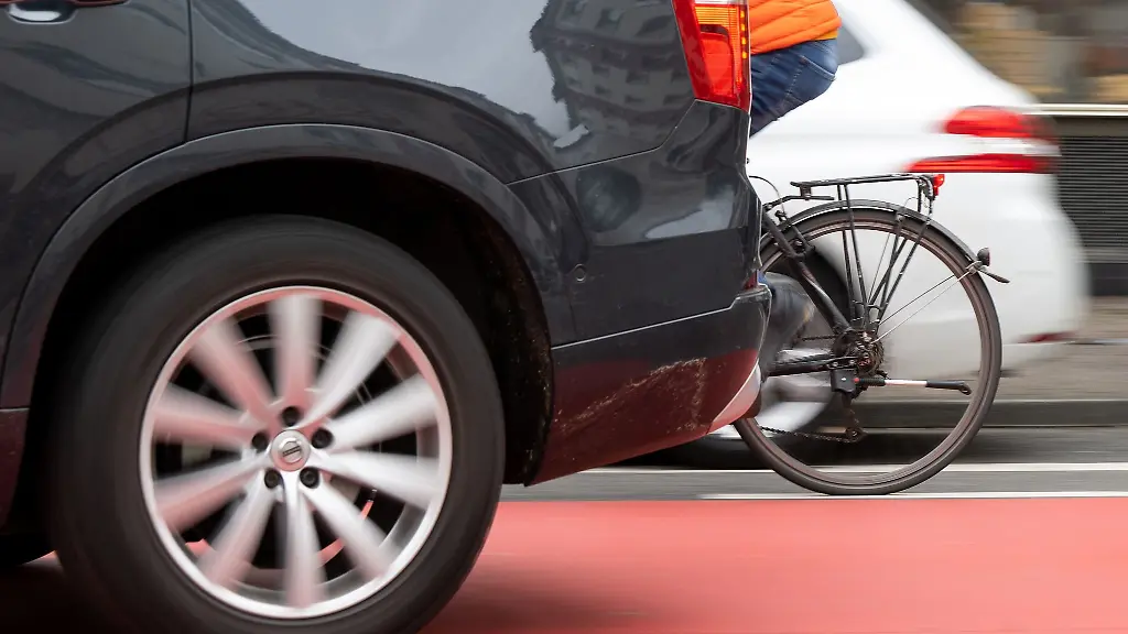 Ein-Fahrradfahrer-faehrt-auf-einer-Fahrradstrasse-zwischen-einem-parkenden-und-fahrenden-Auto-vorbei