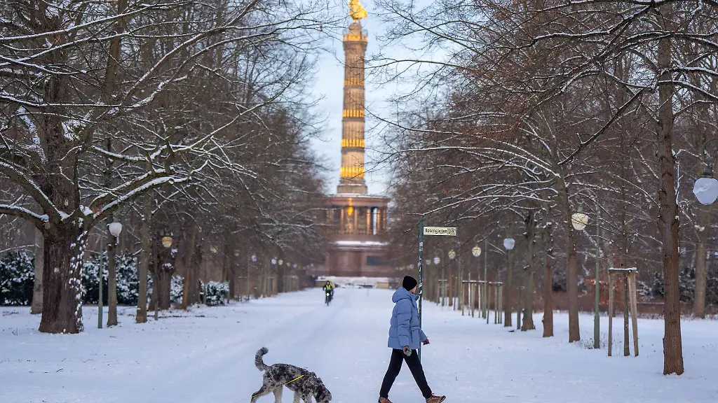 Eine-Frau-geht-mit-ihrem-Hund-vor-der-Kulisse-der-Siegessaeule-im-verschneiten-Tiergarten-spazieren