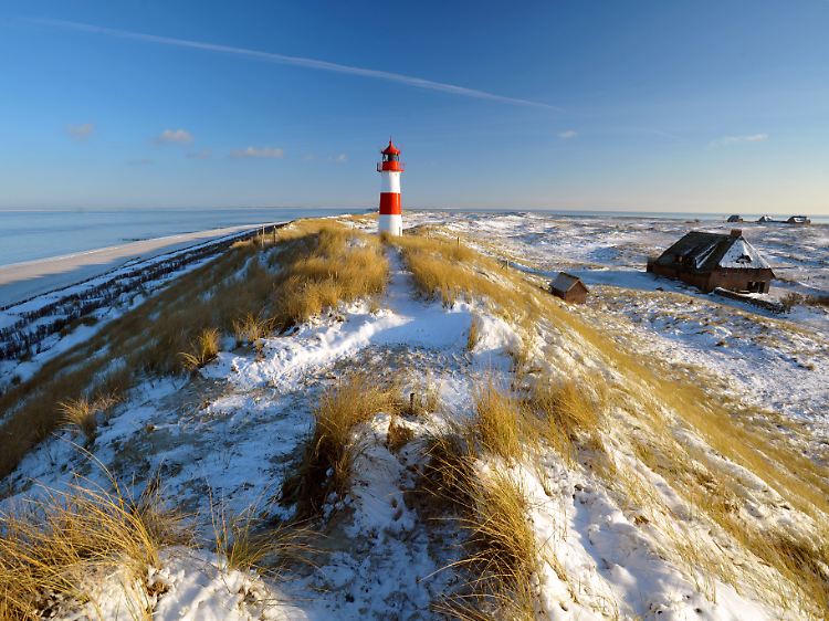 Sylts äußerster Norden im Winter: Reetdachhäuser mit weißen Dächern und der Leuchtturm am Ellenbogen vor verschneiten Dünen.