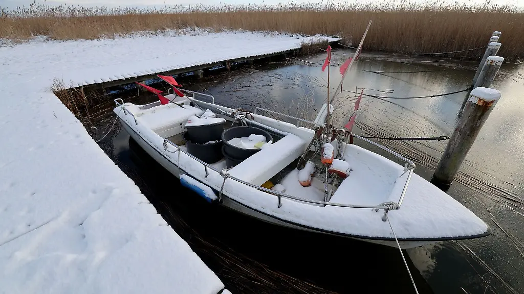 Ein-schneebedecktes-Fischerboot-liegt-im-Boddenhafen-der-teilweise-schon-mit-einer-duennen-Eisschicht-bedeckt-ist