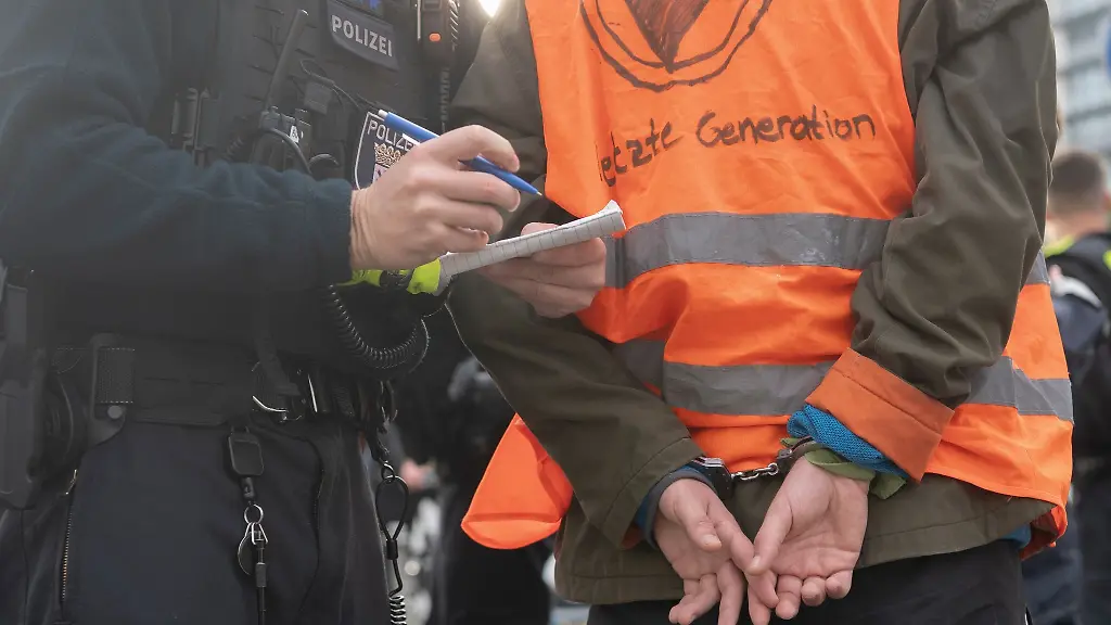 Ein-Aktivist-der-Klimaschutz-Gruppe-Letzte-Generation-steht-in-Handschellen-neben-einem-Polizeibeamten