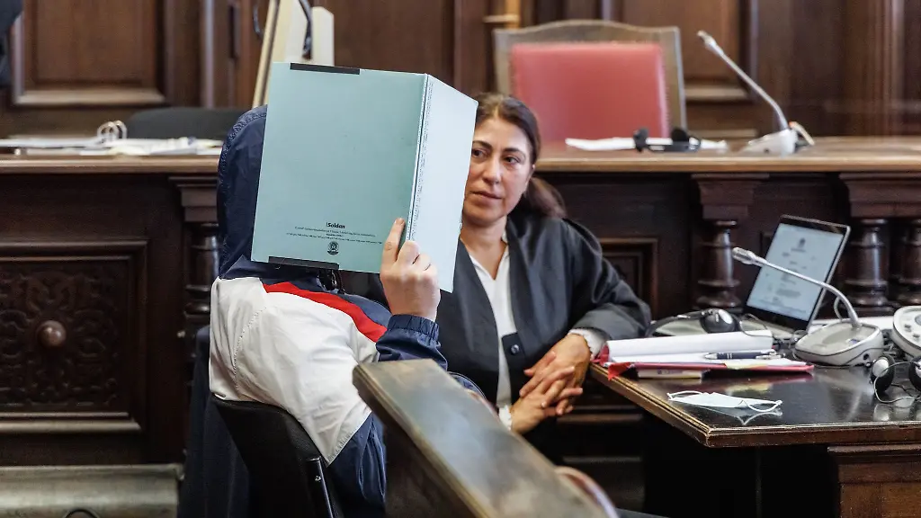Ein-Angeklagter-verbirgt-zu-Prozessbeginn-sein-Gesicht-hinter-einem-Aktenordner