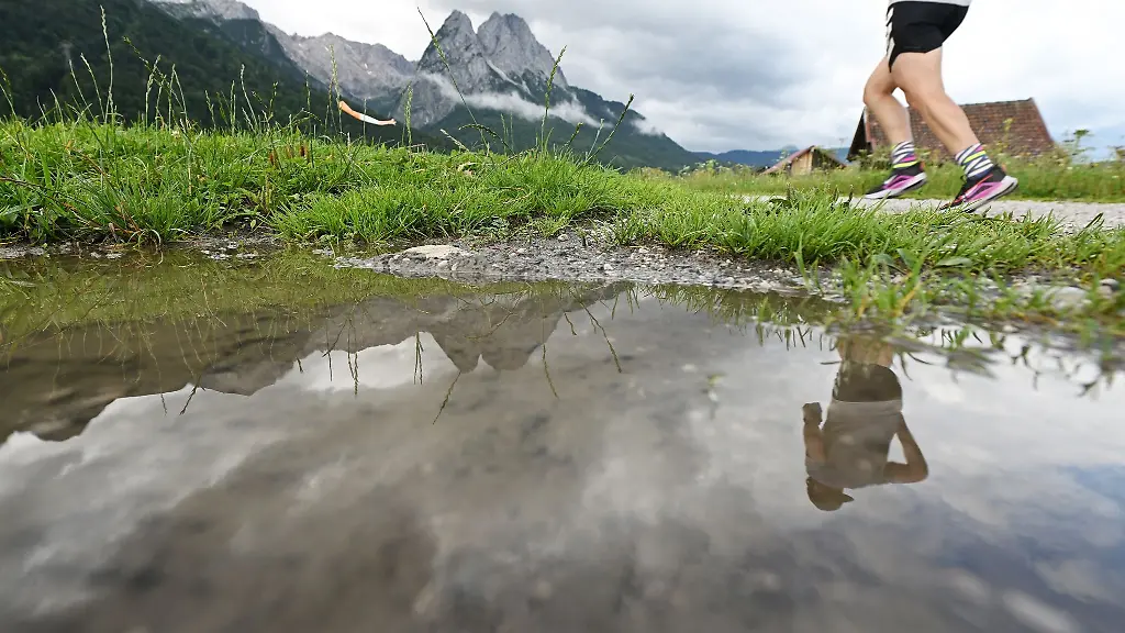 Ein-Jogger-spiegelt-sich-zusammen-mit-den-Gipfeln-des-Wettersteingebirges-in-einer-Regenpfuetze