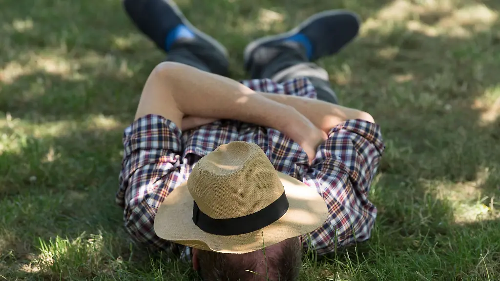 Ein-Mann-mit-einem-Sonnenhut-auf-dem-Gesicht-liegt-auf-einer-Wiese-im-Schatten-und-ruht-sich-aus